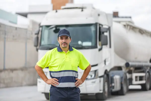 カジュアルなユニフォームとキャップで正のひげ付き男性のメカニカルエンジニアは 道路上のタンカートラックの背景に対して腰に立っている間 カメラを見ています ストック画像