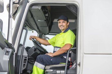 Limonlu ve mavi üniformalı olumlu bir erkek kamyon şoförü açık havada kamyonda oturmuş kameraya bakıp gülümsüyor.