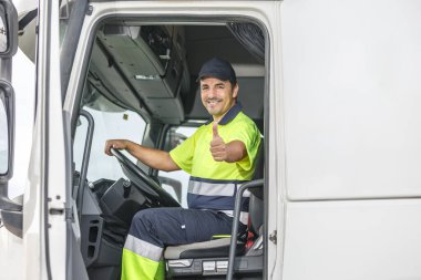 Üniformalı, mutlu, olgun bir erkek sürücü. Gülümsüyor ve jest yapıyor. Kamyonda oturuyor ve yolculuk sırasında kameraya bakıyor.