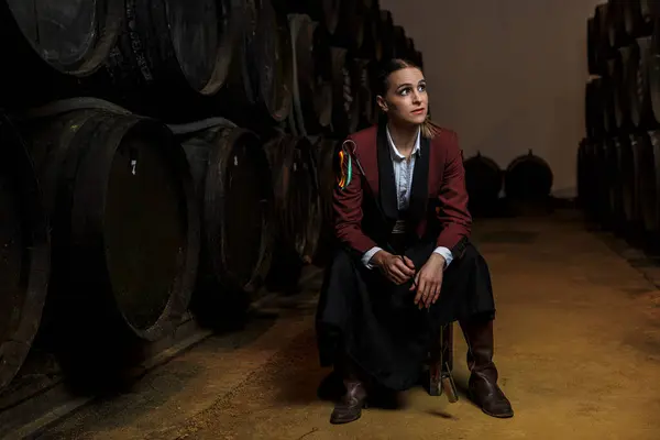 Full body of confident young female venenciador in maroon short coat and maxi black skirt looking up while sitting on chair with venencia in hand and glancing at wine barrels in cellar