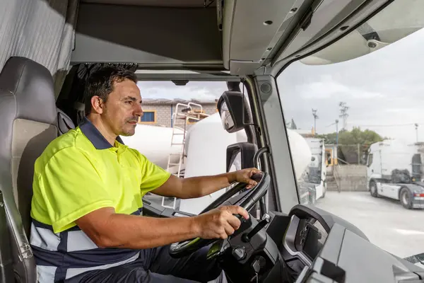 Side view of concentrated middle age male driver in lemon t shirt and curly hair sitting in tanker truck while driving in company compound during work hour