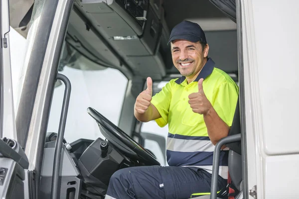ユニフォーム笑顔で陽気な中年男性ドライバーとトラックに座って旅行に出発する前にカメラを見ながら成功した二重親指を示す ロイヤリティフリーのストック写真