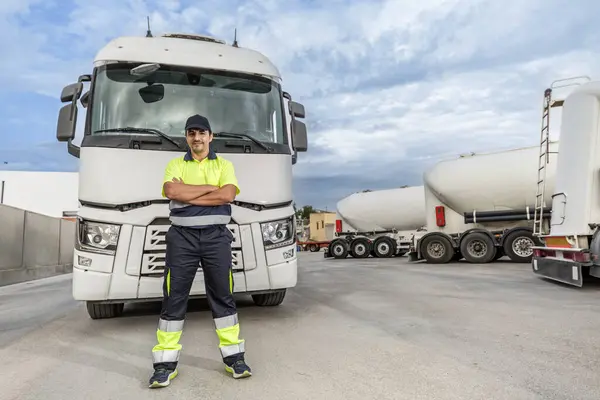 Full body of happy adult male technician in uniform and cap looking at camera while standing with hands crossed in front of large truck against cloudy blue sky and parked vehicles in service center in daylight