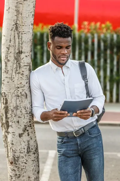 Afro-Amerikalı bir adam bir ağacın yanında duruyor, okul ödevi için bir kitap kullanıyor..