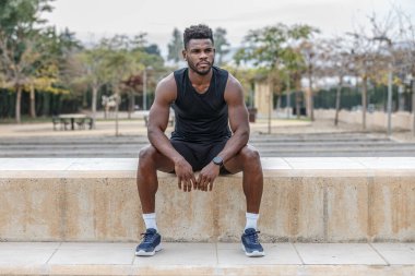 Afrika kökenli güçlü erkek sporcu. Açık hava antrenmanından sonra bulanık ağaçlara karşı oturarak dinleniyor.