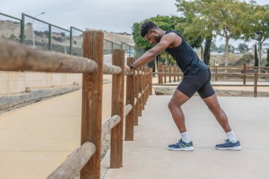 Siyah spor giyim giymiş, tahta çitlere yaslanan ve fitness antrenmanından önce ısınan güçlü Afro-Amerikan sporcunun tüm vücudu yan görünüm.