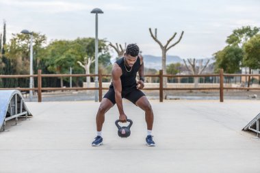 Afrika kökenli Amerikalı erkek sporcu yaz günü şehir parkında antrenman yaparken ağır kettlebell kaldırıyor.