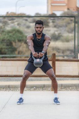 Spor kıyafetleri ve spor ayakkabıları giymiş genç Afrikalı Amerikalı sporcu şehir parkında yoğun eğitim sırasında kettlebell salıncak egzersizi yapıyor.