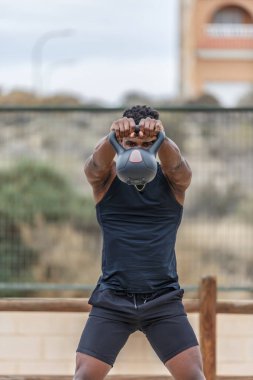Spor kıyafetleri içinde genç Afrikalı Amerikalı sporcu şehir parkında antrenman yaparken ağırlık kaldırıyor gündüz vakti kettlebell salıncak egzersizi yapıyor.
