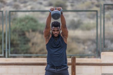 Aktif giyimli, Afro saçlı, spor sahasında çitlere karşı yoğun spor yaparken kettlebell salıncak egzersizi yapan güçlü siyah bir sporcu.