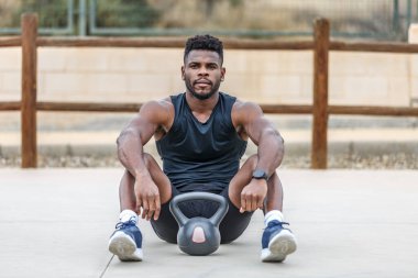 Sakallı, Afro-Amerikan spor giyim sporunda kettlebell ile oturmuş kameraya bakan genç ve kaslı bir sporcu.