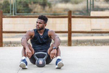Spor kıyafetleri ve spor ayakkabıları giyen kararlı etnik sporculardan oluşan bir vücut. Açık hava fitness antrenmanı sırasında elinde kettlebell ile yerde oturuyor ve başka tarafa bakıyor.