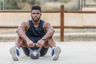 Spor ayakkabılı sakallı sporcu ve spor çanlı aktif giyimli spor ayakkabısı spor sahasında, spor yaparken kameraya bakarken bulanık arka planda oturuyor.