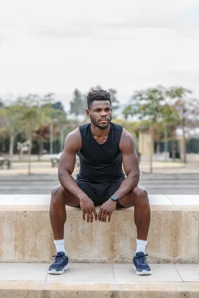 Full body of African American male athlete in sportswear sitting on border during fitness training and resting