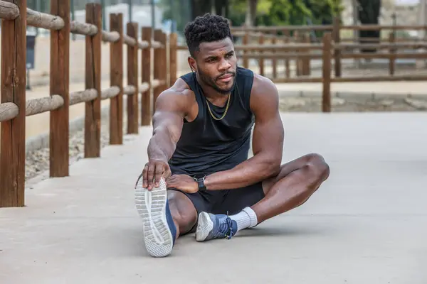 Full body of young strong African American runner sitting on ground and stretching leg muscles while pulling toes before training