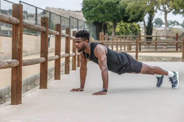 Full body of strong African American male athlete performing plank exercise on ground near wooden fence during fitness workout