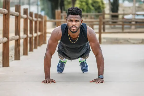 Full body of strong African American sportsman with Afro hair doing plank exercise near wooden fence during fitness workout in park