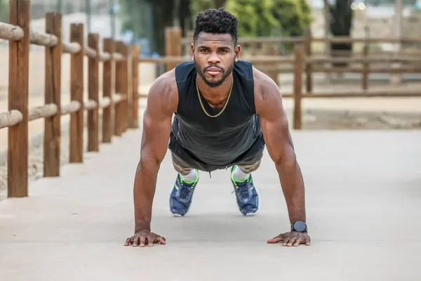 Full body of concentrated young African American athlete with muscular arms doing plank exercise on ground during fitness workout in summer park