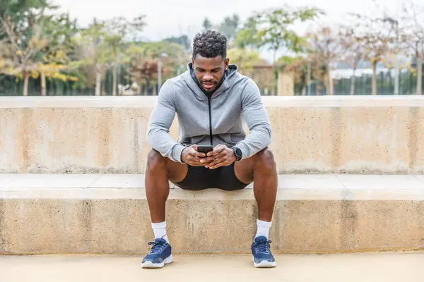 Full body of young African American male athlete in activewear sitting on stairs and messaging via mobile phone during break from outdoor training