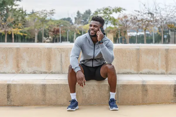 Full body of happy young African American athlete in activewear sitting on stairs and talking via mobile phone during break from workout