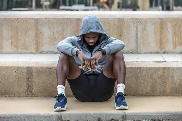 Full body of young tired and melancholic African American male athlete with hood on head sitting on stairs after outdoor training