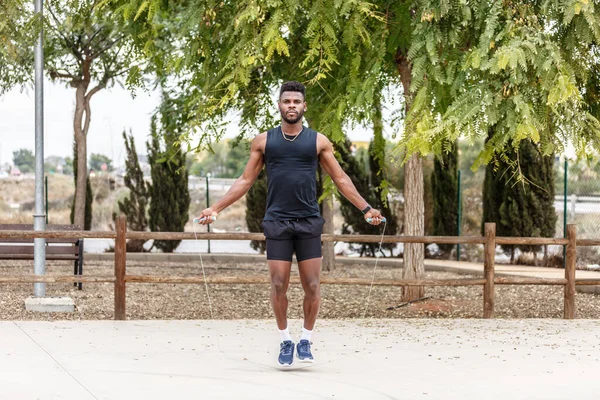 Full body of determined African American male athlete in sports outfit and sneakers jumping rope in summer park during cardio training
