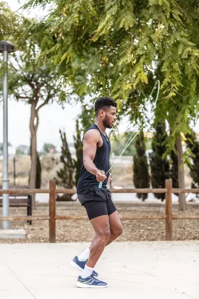 Side view full body of young African American athlete in sports outfit training with jumping rope near green trees during fitness workout