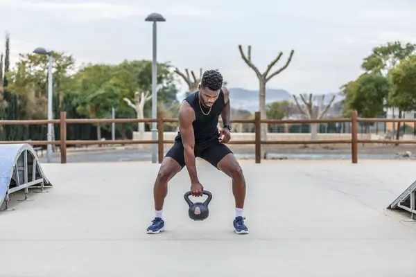 Full body of determined African American male athlete lifting heavy kettlebell while training in city park on summer day