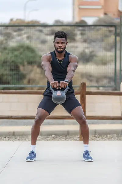 Full body of young African American athlete in sports clothes and sneakers doing kettlebell swing exercise during intense training in city park