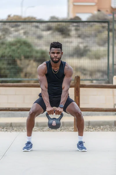 Full body of determined African American male athlete with Afro hair training with heavy kettlebell on sports ground in city