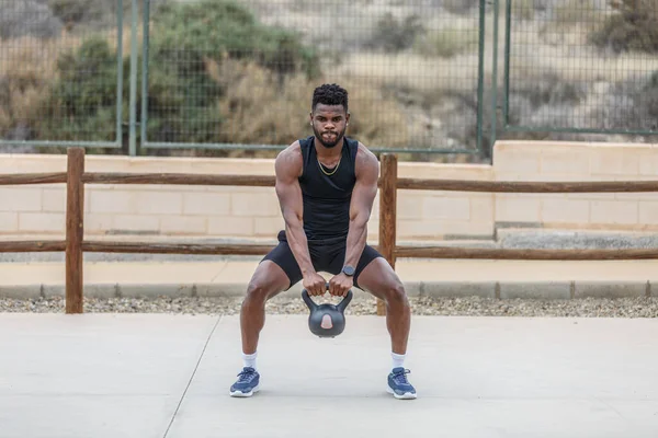 Full body of determined African American male athlete doing squats with heavy kettlebell during intense training on sports ground