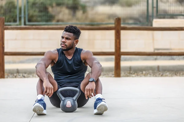 Full body of determined ethnic athlete in sportswear and sneakers sitting on floor with kettlebell during outdoor fitness training and looking away