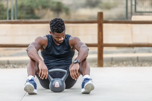Full body of determined African American sportsman resting on ground with heavy kettlebell after intense weightlifting training in daylight