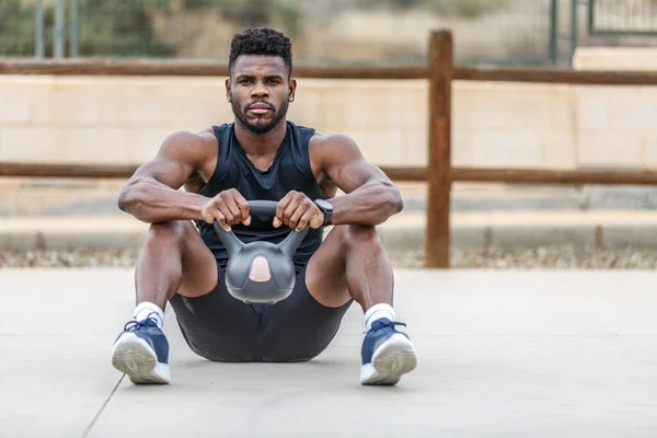 Full body of confident bearded athlete in sportswear and sneakers sitting and lifting heavy kettlebell during outdoor weightlifting training on summer day