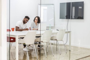 Bir adam ve bir kadın modern bir ofiste beyaz bir konferans masasında bir proje üzerinde çalışıyorlar. Kadın çizgili gömlek ve gözlük takarken adam beyaz bir gömlek ve gözlük takıyor. İkisi de masadaki bir belgeye bakıyor. Th