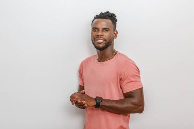 Genç bir adam sıcak bir gülümseme ile, rahat pembe bir tişört giyerek güvenle ayakta duruyor. Elleri önünde birbirine kenetlenmiş ve bileğinde modaya uygun bir saat var. Tarafsız gri arkaplan, onun sakin tavrını ve dostça ifadesini vurguluyor.