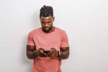 Gündelik pembe tişört giyen genç bir adam akıllı telefonuna dikkatle odaklanıyor, aktivitelere dalmış. Saç stili kısa, bukleli ve arka plan da basit, nötr bir renk. Bu da cihaza yoğunlaşmasını sağlıyor..