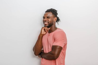 Şık bir saç stili olan genç bir adam geniş bir gülümseme içinde, çenesini elinin üzerinde dinlendiriyor. Açık renkli bir duvara yaslanmış rahat bir tavır ve kendinden emin bir ifade sergiliyor. Ayarlama bir pozitiflik ve yaklaşım hissi veriyor.