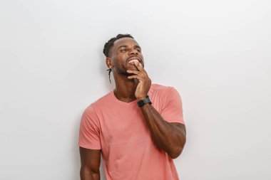 Açık pembe tişört giyen genç bir adam düz bir duvara yaslanmış gülümsüyor ve düşünceli bir şekilde çenesine dokunuyor. Rahat duruşu ve kendine güveni çağdaş bir iç mekanda bir anlık tefekkür ya da eğlence olduğunu gösteriyor..