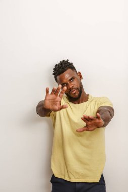 Bir adam açık renkli düz bir duvara karşı duruyor, kollarını şakacı bir şekilde öne uzatıyor. Sarı tişört ve siyah pantolon giyerken rahat bir tavır sergiliyor, etkileyici ve kendinden emin bir vücut dili sergiliyor..