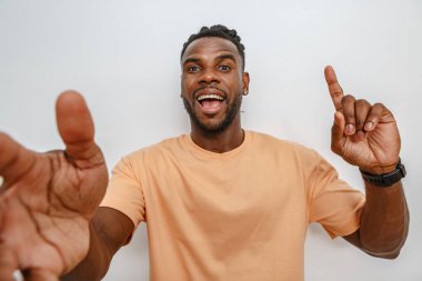 Geniş tebessümlü genç bir adam kollarını yukarıyı göstererek sevinçle uzatıyor. Açık şeftali rengi bir gömlek giymiş ve rahat bir saç stiline sahip. Basit bir arka plana karşı heyecan ve pozitiflik ifadesi sergiliyor..