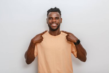 Bir adam rahat bir şeftali gömlek giyerken kendine güvenerek, geniş bir gülümsemeyle ayakta duruyor. Kendini şakacı bir şekilde gösteriyor, sade, tarafsız bir arkaplana karşı neşeli bir yüz ifadesi sergiliyor, canlandırıcı bir anı yakalıyor..