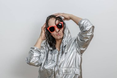 Islak saçlı genç bir kadın parlak gümüş yağmurluk ve parlak kırmızı güneş gözlüğü takarken neşeli bir poz verir. Saçını şakayla düzeltiyor, minimalist bir ortamda kendine güvenen ve kaygısız bir tavır sergiliyor..