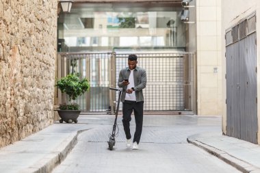 A casually dressed African-American man rides an electric scooter down a street while consulting his smartphone, heading towards a coworking center, showing a mix of work and mobility. clipart