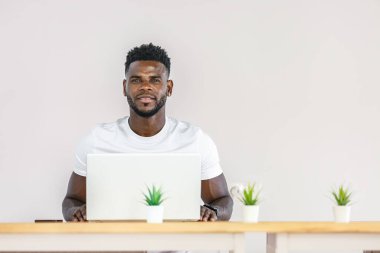 A young man sits at a wooden desk focusing on his laptop, surrounded by small potted plants in a clean, modern workspace, emphasizing a productive and serene environment. clipart