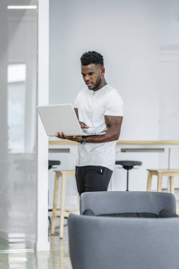 Genç bir adam, çağdaş bir ofiste dizüstü bilgisayarına odaklanırken ayakta duruyor. Parlak çevrede şık mobilyalar ve iş için üretken bir atmosfer yer alıyor..
