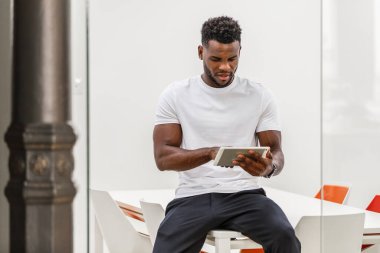 30 yaşında, Afro-Amerikan bir adam, parlak, çağdaş bir ofis masasında oturuyor. Teknoloji ve yaratıcılığın bir karışımını sergilemek için bir tablet kullanmaya odaklanmış..