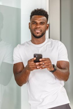 Bir adam akıllı telefonuna odaklanmış, parlak cam bir duvara yaslanmış. Düz beyaz bir tişört giyiyor ve kendine güvenen bir ifadesi var, modern, kentsel bir yaşam tarzını somutlaştırıyor..