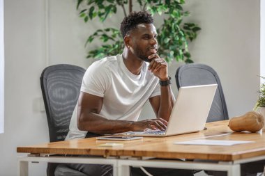 Bir adam, dizüstü bilgisayarı üzerinde çalışırken derin düşüncelere dalmış, parlak bir ofisin ahşap masasında oturuyor. Bereketli yeşillik çalışma alanı atmosferine ferahlatıcı bir dokunuş ekler.