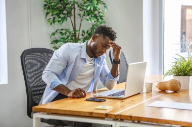 Çağdaş bir ofis ortamında, bir adam tahta masasında otururken stresli görünür. Alnını açık bir şekilde endişeli tutuyor. Etrafı yeşilliklerle çevrili laptopuyla çalışıyor..
